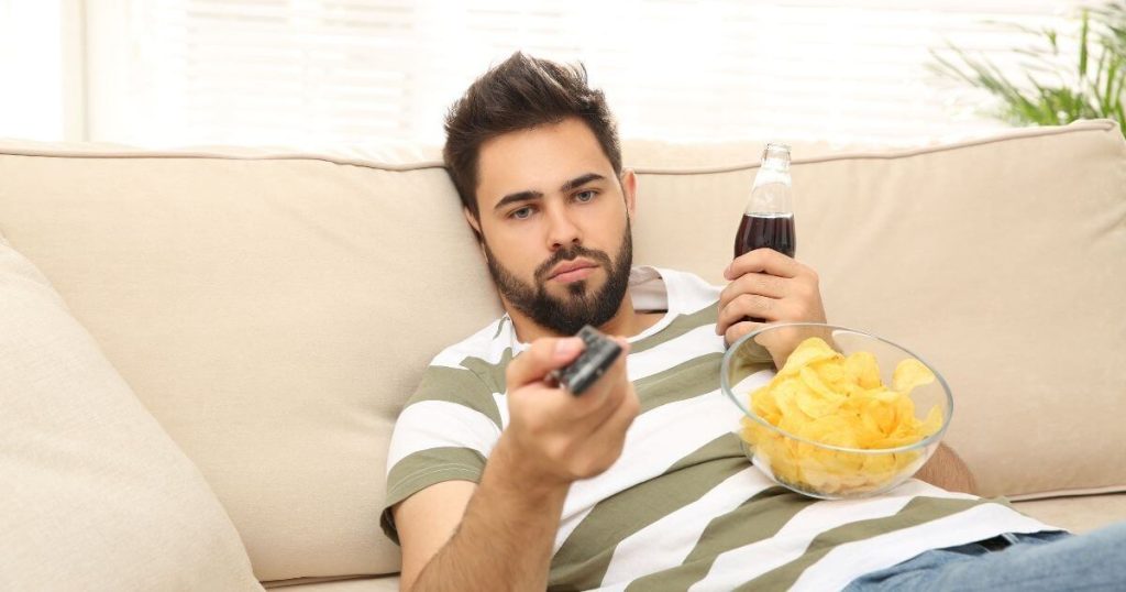 Man relaxing on a sofa with snacks and a drink, representing a sedentary lifestyle